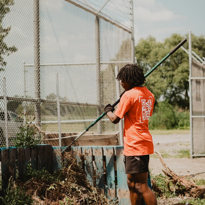 A young Black person in an orange shirt uses a rake to turn a large compost pile in a wooden bin next to a tall chain-link fence on a sunny day.